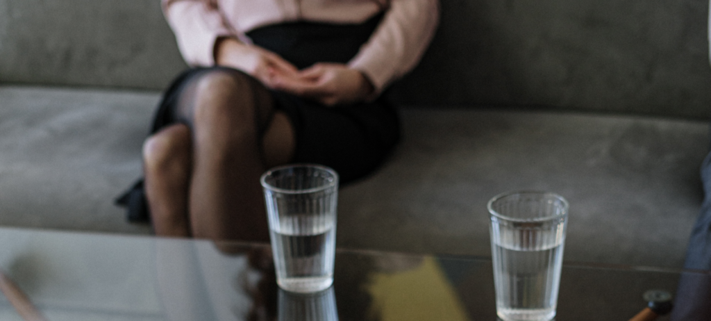 Dame siddende i baggrunden på en grå sofa, vi ser hænderne liggende i skødet og knæene. FOran er et bord med to glas med vand i.