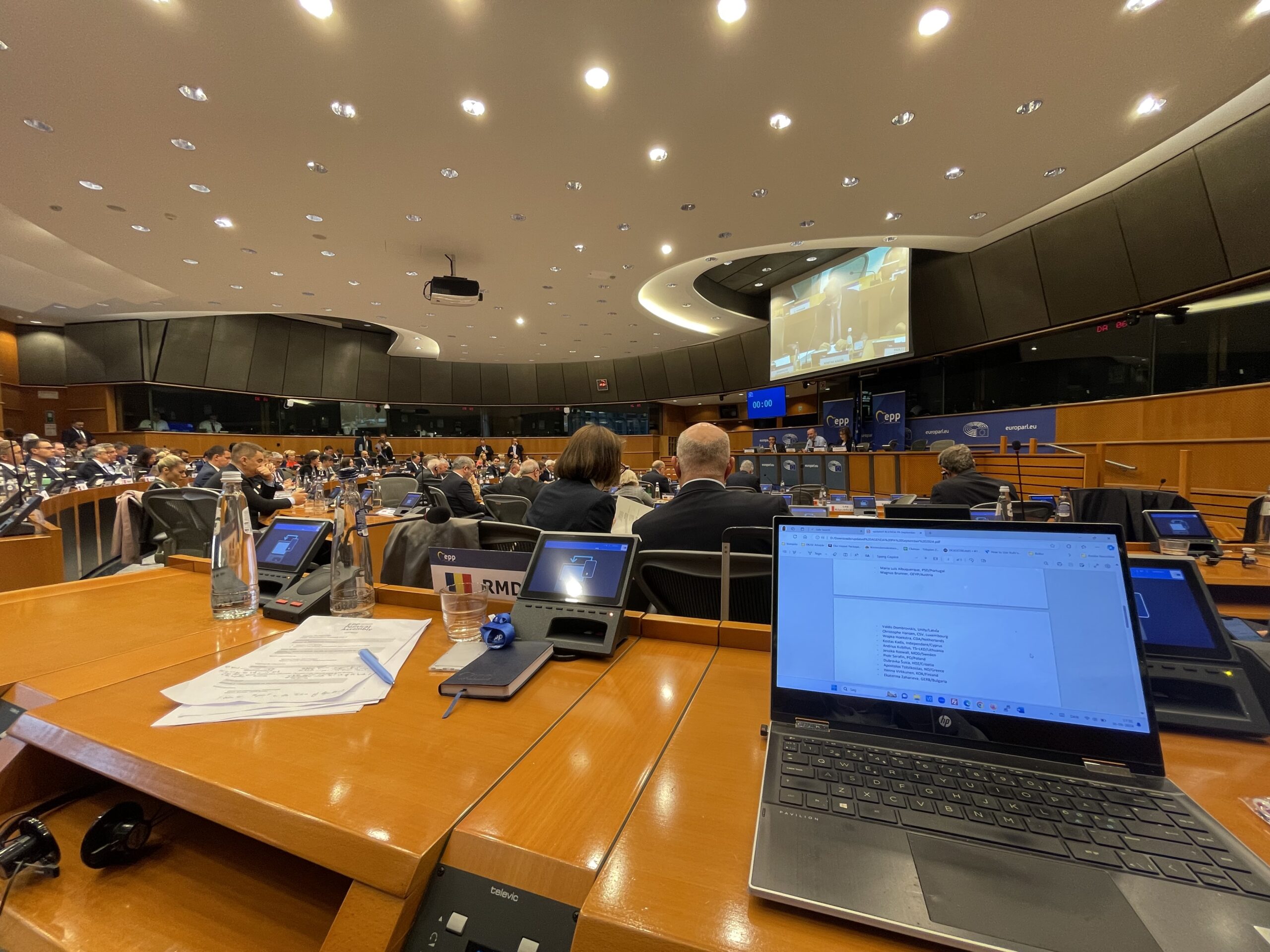 Oversigt over plenarsalen i Europa-Parlamentet i Bruxelles under EPP Political Assembly. I forgrunden ses borde med b&aelig;rbare computere, dokumenter og navneskilte, herunder et rum&aelig;nsk flag og RMDC-skiltet. I baggrunden ses deltagere ved bordene med EPP-logoer og sk&aelig;rme synlige p&aring; podiet.