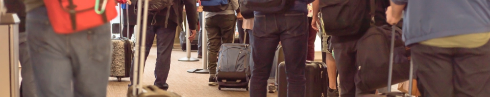 Mennesker med kufferter og bagage i k&oslash; i en lufthavn eller gr&aelig;nseovergang, fotograferet fra siden i &oslash;jenh&oslash;jde. Kun benene og bagagen er synlige.