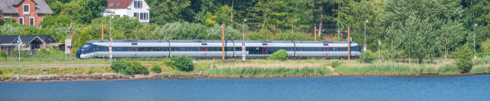 Et DSB IC4-tog k&oslash;rer langs en dansk fjord omgivet af gr&oslash;n natur og danske huse i baggrunden. Billedet er taget fra den modsatte bred af fjorden p&aring; en solrig sommerdag.