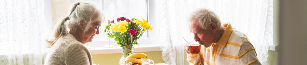 Et &aelig;ldre &aelig;gtepar sidder sammen ved et bord ved et lyst vindue med gardiner. Manden drikker te mens kvinden ser p&aring; ham med et smil. P&aring; bordet st&aring;r en buket med gule og r&oslash;de blomster samt frugt. Stemningen er varm og n&aelig;rv&aelig;rende.