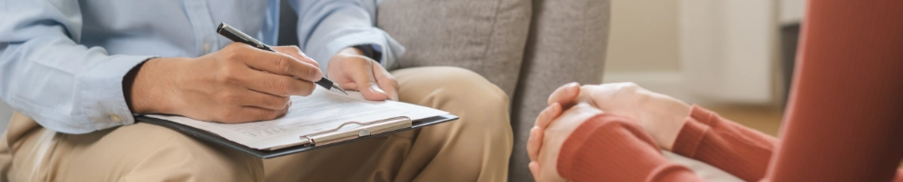 N&aelig;rbillede af en sundhedsfaglig person i lysebl&aring; skjorte der skriver p&aring; en clipboard, mens en patient med foldede h&aelig;nder sidder overfor i en sofa. Kun h&aelig;nder og overkroppe er synlige.